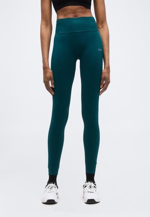 Persoană cu abdomen tonifiat, purtând colanți cu talie înaltă de culoare teal și adidași albi, stând pe un fundal simplu.