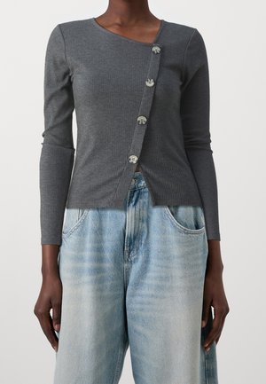 Personne à la peau foncée portant un haut ajusté gris charbon à manches longues côtelé avec un design asymétrique à boutons sur le devant et un jean large bleu clair.