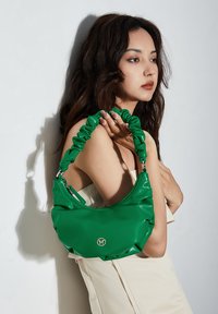 Nainen, jolla on pitkät tummat hiukset, pitää rypistyneenä vihreää käsilaukkua olkapäällä, pukeutuneena ilman hihoja olevaan beigeen mekkoon yksinkertaista taustaa vasten.