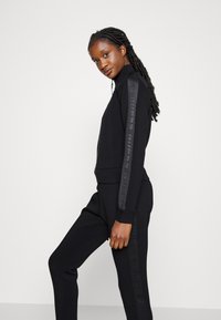 Zwarte rits-sweater en bijpassende broek, gemaakt van gladde stof. Heeft een hoge kraag en logo-accenten langs de mouwen.