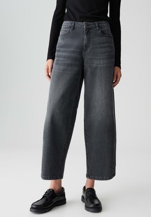 Personne portant un jean large taille haute gris foncé et des chaussures noires à lacets, se tenant devant un fond clair uni.