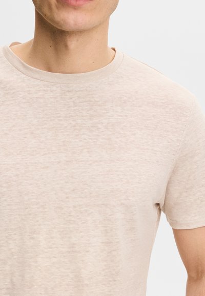 Homme portant un t-shirt beige clair à manches courtes avec col rond sur fond blanc uni, recadré au niveau de la bouche et du milieu du bras.