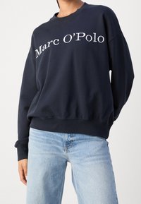 Granatowa bawełniana bluza z logo "Marc O'Polo" w białym skrypcie. Projekt zawiera okrągły dekolt oraz ściągacze przy rękawach, z luźnym krojem.