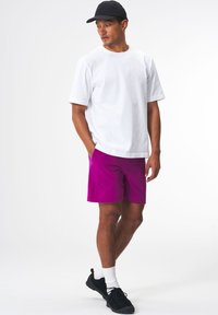 Weißes Baumwoll-T-Shirt und lila Shorts. Das T-Shirt hat einen Rundhalsausschnitt; die Shorts verfügen über seitliche Taschen und einen elastischen Bund. Schwarze Sportschuhe und eine Cap.