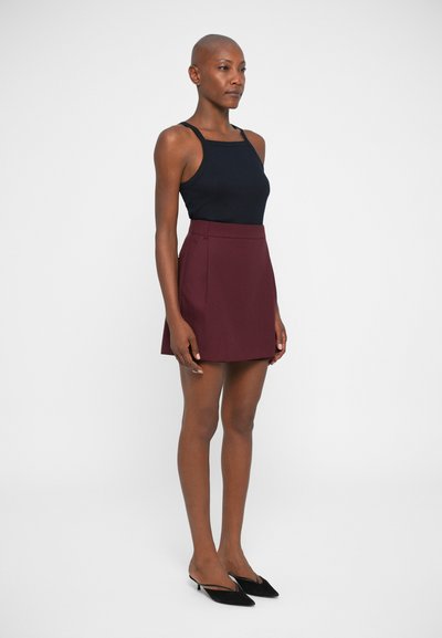 Débardeur noir à bretelles spaghetti associé à une jupe bordeaux au-dessus du genou. La jupe présente un design épuré et des détails de plis. Mules noires à talons.