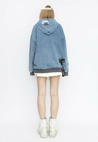 Sudadera oversized de denim azul con capucha, que presenta una etiqueta de logo y acentos en gris contrastante. Llevada sobre una minifalda blanca y unas botas contundentes blancas.