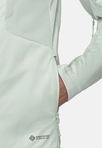 Hellgrüne Softshelljacke mit Reißverschlusstasche, elastischen Bündchen und dem Logo "WINDSTOPPER by GORE-TEX LABS". Strukturiertes Stofffinish.