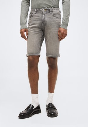 Homme portant un short en jean gris, une chemise grise à manches longues, des chaussettes blanches et des mocassins en cuir noir, debout devant un fond uni.