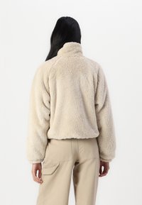 Beżowa, puszysta kurtka z wysokim kołnierzem o luźnym kroju i długich rękawach. Miękka faktura z krótkim fasonem, zestawiona z jasnymi spodniami.