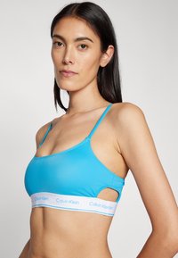 Šviesiai mėlynas braletas, pagamintas iš lygios medžiagos, su plonomis petnešomis ir iškirpimo detalėmis. Yra balta juosta su "Calvin Klein" logotipu.