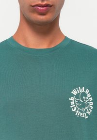 Kratak izrez osobe koja nosi zelenu majicu s bijelim grafičkim prikazom crtanog pačina i natpisom "Wild Runners Trailster Club" na prsima.