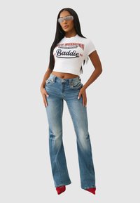 Biała krótka kurtka T-shirt z czerwonym i niebieskim napisem, zestawiona z jasnoniebieskimi dzwonami. Przezroczyste okulary przeciwsłoneczne i czerwone szpilki dopełniają outfit.