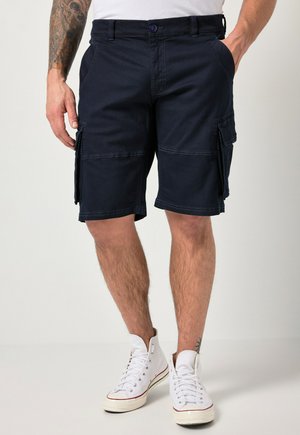 Mann trägt dunkelblaue Cargo-Shorts mit Seitentaschen, weiße hohe Sneaker und ein weißes Hemd, steht vor einem schlichten Hintergrund.