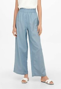 Person som bär ljusblå vida byxor med elastisk midja, vit ärmlös topp och vita öppna sandaler på en vit bakgrund.