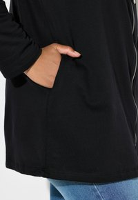 Giacca nera a maniche lunghe con cerniera frontale, dotata di tasca laterale e orlo dritto, realizzata in tessuto morbido ed elastico.