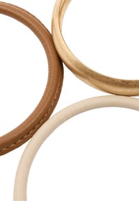 Trzy bransoletki: brązowy pasek ze skóry z przeszyciami, gładki kremowy pasek ze skóry oraz błyszczący złoty pasek metalowy, zachodzące na siebie.