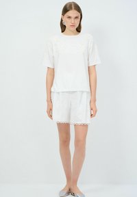 Camiseta blanca de algodón con adornos detallados en los hombros, combinada con pantalones cortos bordados a juego con encaje en los bordes. Bailarinas plateadas.
