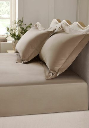 Beiges Bett mit passenden Kissenbezügen und Spannbettlaken, wellenförmiges gepolstertes Kopfteil und eine Vase mit Blumen auf einem nahen Tisch am Fenster.