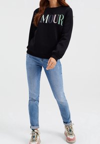 Svart sweatshirt med "AMOUR" i pastellfärger, kombinerad med ljusblå jeans och flerfärgade sneakers. Avslappnad, figursydd design.