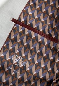 Tela gris con un patrón geométrico en marrón, morado y crema. Presenta una etiqueta que dice "NEXT ATHLETIC FIT 40R" y un ribete en burdeos.