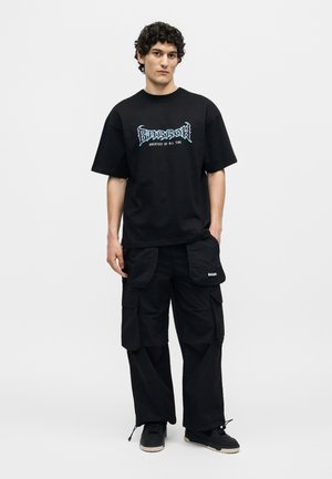 Giovane uomo in piedi con t-shirt nera con stampa grafica e scritta “BARROW”, pantaloni cargo neri con molte tasche e sneaker nere.