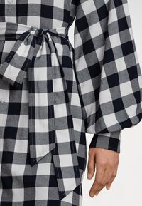 Melns un balts rūtainais kleitas ar garām piedurknēm, kas saistīta ar jostas siksnu viduklī, un redzama roka blakus gurnam.