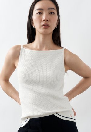 Ženska z dolgimi temnimi lasmi, oblečena v belo teksturirano brezrokavnik in črne hlače, stoji z rokami na bokih pred enobarvnim ozadjem.