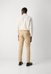 Man staat met zijn rug naar de camera gekeerd, in een crème shirt, beige broek en zwarte schoenen, tegen een effen witte achtergrond.