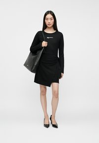 Naine kannab musta pika varrukaga kleiti ja musti kontsasid, hoides õlal suurt musta nahast kott.