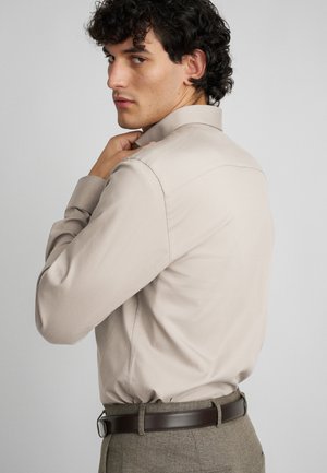 Camisa bege de mangas longas com colarinho clássico, apresentando tecido suave e um corte ajustado, combinada com um cinto castanho e calças cinzas texturizadas.