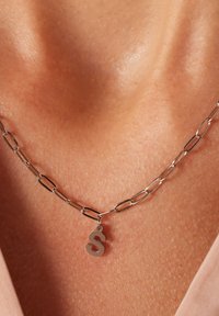Zilveren ketting met ovale schakel, voorzien van een kleine "S" hanger. Gladde metalen oppervlakte, close-up zicht tegen de huid.