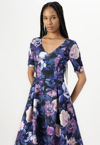 Virágmintás ruha V-kivágással, rövid ujjakkal és A vonalú sziluettel. Az anyagon nagy lila és rózsaszín virágok találhatók sötét háttéren.
