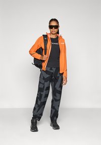 Oranges wasserdichte Jacke, schwarzes T-Shirt mit Grafik, gemusterte schwarze Hose und schwarze Sneaker. Accessoires sind Sonnenbrille und ein schwarzer Rucksack.