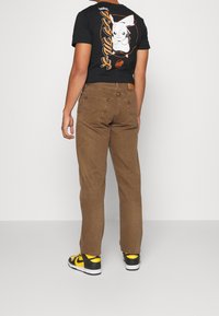 Bruna raka jeans i kombination med en svart t-shirt med grafik, som har en Pikachu-design, bärs tillsammans med svart och gula sneakers.