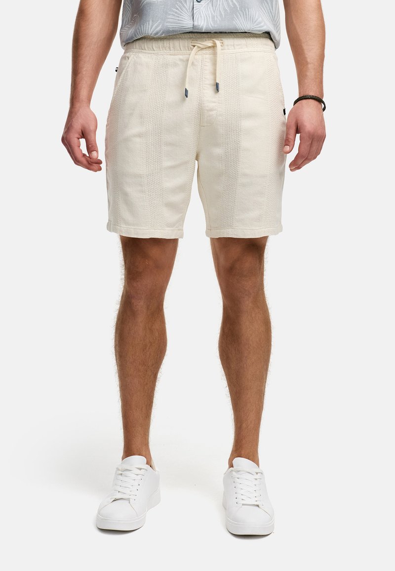 Helle beige Shorts aus strukturiertem Stoff mit einem Kordelzugbund. Verfügt über Seitentaschen und umschlagbare Säume. Getragen mit weißen Sneakers.