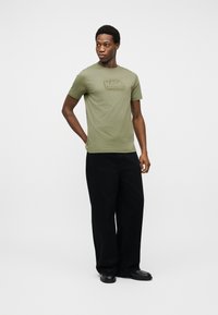 Jeune homme debout portant un t-shirt olive de Karl Lagerfeld, un pantalon noir ample et des chaussures noires, sur un fond clair uni.