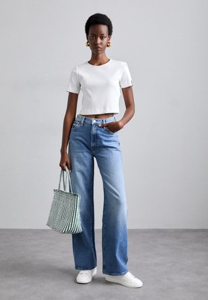 T-shirt cropped branca com textura canelada, jeans de perna larga azul-claro e ténis brancos. A segurar uma mala de ombro riscada em verde e branco.