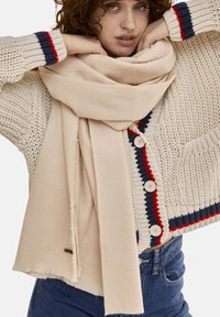 Beige tørklæde lavet af blødt stof, draperet afslappet. Det har frynser i kanten. Modellen bærer en strikket sweater med multikolor accentuer.