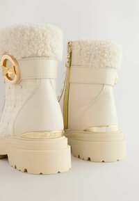 Witte leren enkellaarzen met pluche crèmekleurige bontmanchetten, gouden hardwareaccenten, een decoratieve gesp en getextureerde blokhakken.