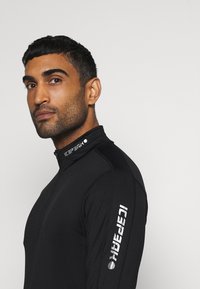 Haut de sport zippé noir avec col haut. Caractérisé par le logo argent "ICE PARK" sur le col et la manche gauche. Texture lisse, design ajusté.