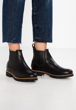 Botas Chelsea de cuero negro con paneles laterales elásticos, suelas de goma gruesa y un ribete de cuero marrón, llevadas con unos vaqueros cortos azules.