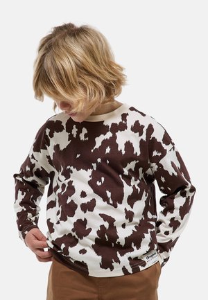 Enfant aux cheveux blonds portant un t-shirt à manches longues imprimé tache de vache marron et blanc ainsi qu'un pantalon marron, regardant vers le bas et tenant l'ourlet du t-shirt.