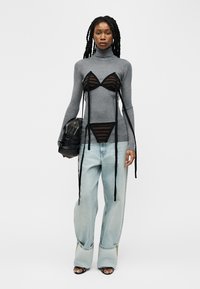 Γυναίκα με μακριά πλεξούδες φοράει γκρι πουλόβερ με ζιβάγκο, μαύρο διάφανο ριγωτό σουτιέν και εσώρουχα από πάνω, άνετο ανοιχτόχρωμο τζιν, κρατώντας μαύρη τσάντα.