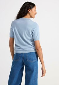 Lys blå strikket top med korte ærmer og rund halsudskæring, parret med højtaljede blå denimjeans med to baglommer.