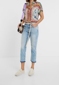 Camisa de manga corta con estampado colorido y detalles en verde, combinado con unos pantalones vaqueros de mezclilla azul claro con dobladillos deshilachados y un detalle bordado. Zapatillas beige.