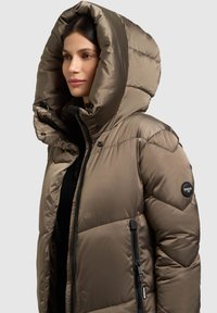 Beeziline beež paksenisti mantel suure ülikattega kapuutsiga, varustatud tõmblukuga küljetaskuga ja brändi logoga. Materjal on läikiva pinnaga ja tikitud mustriga.