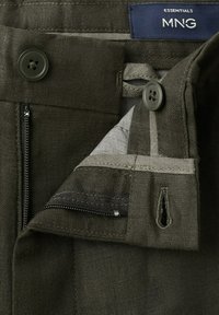 Pantalon vert foncé avec fermeture à boutons et zip, montrant les détails des coutures intérieures et une étiquette bleue portant l'inscription "ESSENTIALS MNG".