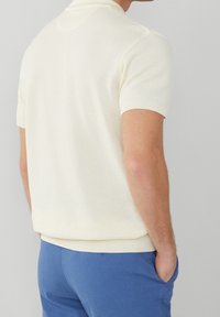 Ljuskrämfärgad kortärmad polo med en texturerad stickad design och ribbad krage. Buren tillsammans med blå byxor, vilket visar en åtsittande silhuett.