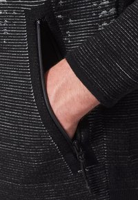 Hand inserted into the zippered pocket of a black and gray striped textured jacket sleeve.