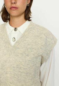 Világosszürke kötött mellény V-nyakkal, rövid ujjakkal és puha textúrával, fehér galléros ing felett, dekoratív gombokkal.